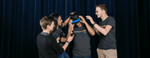 One student uses a virtual reality headset while three other students assist him.