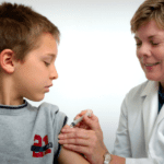 Young boy with blond hair looks on as a doctor with short blonde hair gives him a shot on the arm.