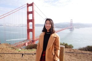 Woman with shoulder length dark brown hair wearing a tan coat stands in front of the golden gate bridge.